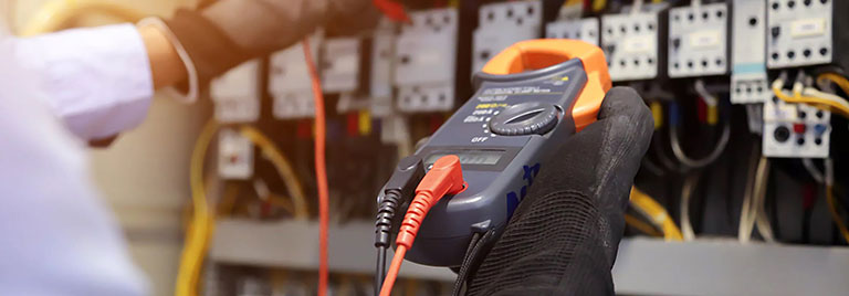 A person wearing gloves uses a digital multimeter to test electrical circuits in a control panel.